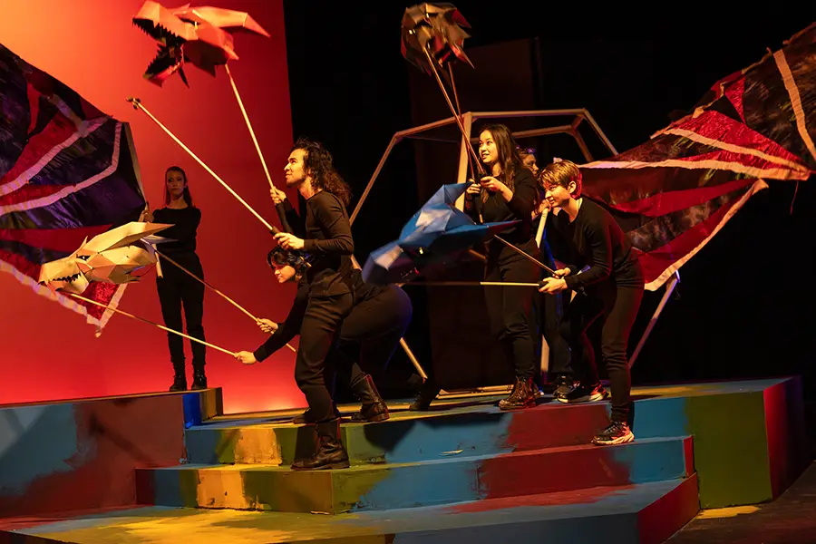 A group of students rehearses on stage while all dressed in black, and holding props of dragon heads.