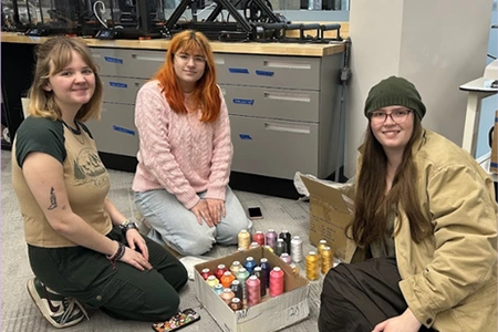 3 students sit on the floor with spools of thread 