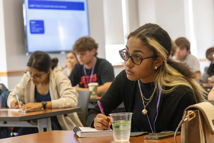 A student sits a table taking notes. Other students are at tables behind her.