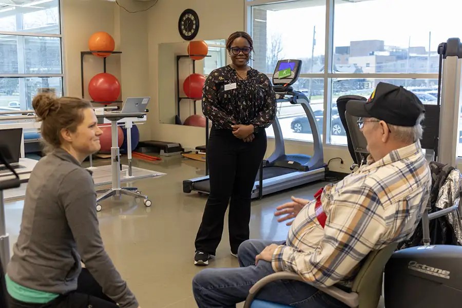 A student dressed in professional clothing, stands between two people. One, an elderly person in a wheelchair, and the second looks to be sitting on an exercise ball.