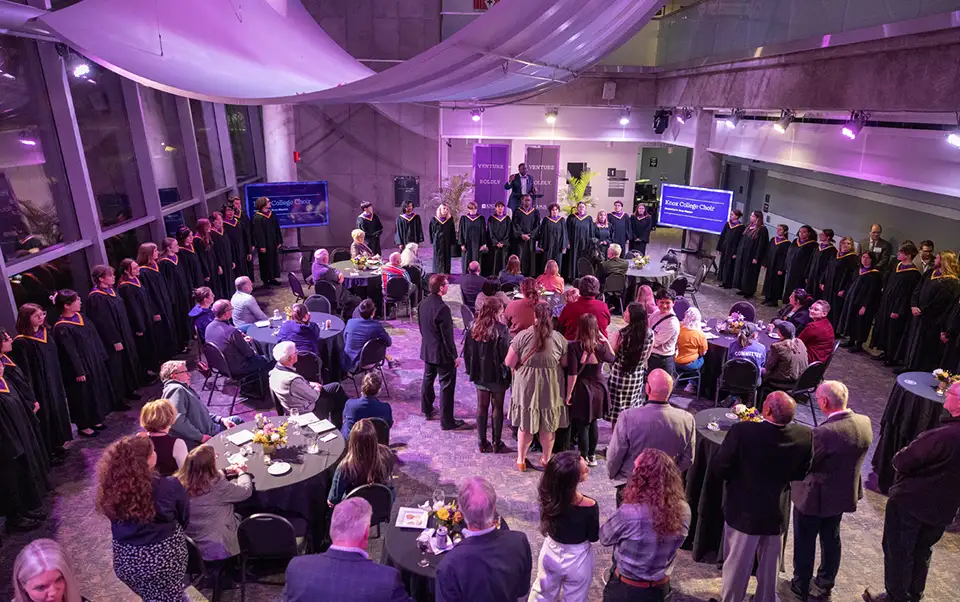 A large group of guests stand in the middle of the room, with the stage at the front of them, and the choir lining around the room. 