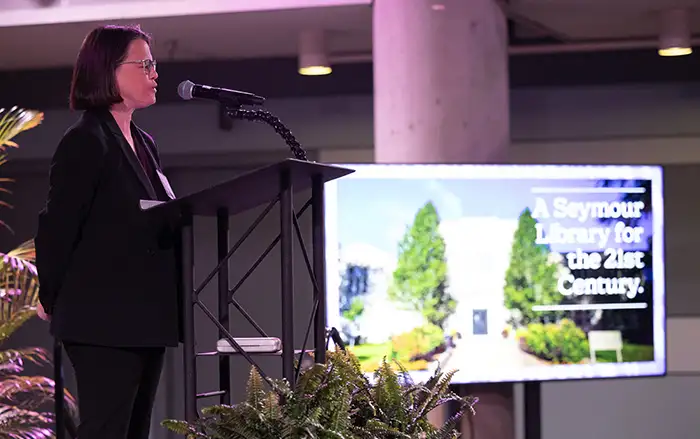 Anne Thomason stands at the podium speaking to the crowd during Venture Boldly in Denver. A monitor can be seen in the background displaying the Seymour Library.