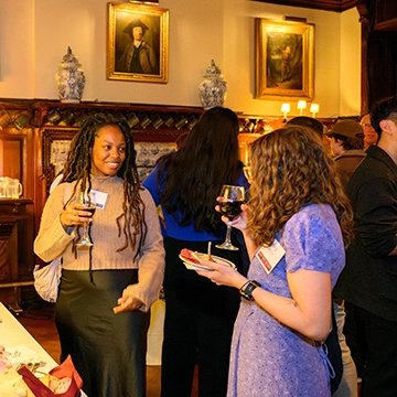Two alums stand while holding drinks, talking and laughing with each other. 
