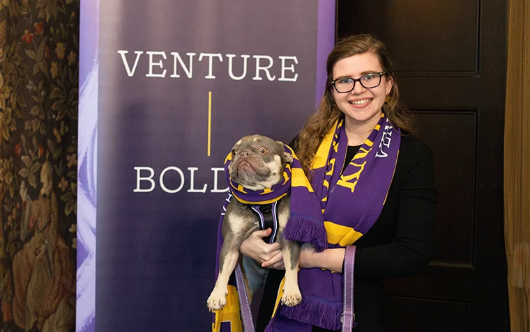 A woman holds a dog and smiles in front of a purple Venture Boldly banners. Both the dog and the woman are wearing a Venture Boldly scarf.