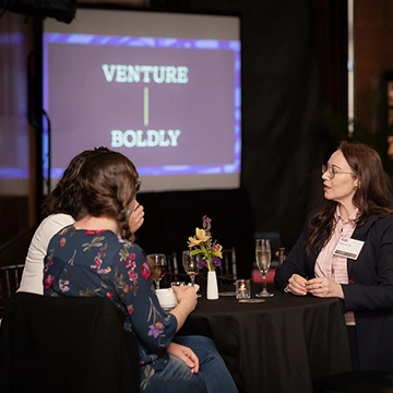 Three event attendees converse at a table; text "Venture Boldly" appears on a screen behind them.
