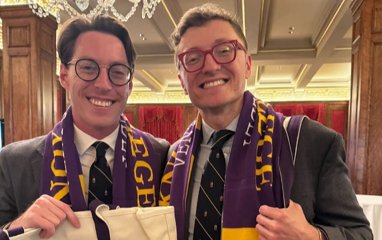 Two men wearing Venture Boldly scarves and holding Venture Boldly Tote Bags smile at the camera.
