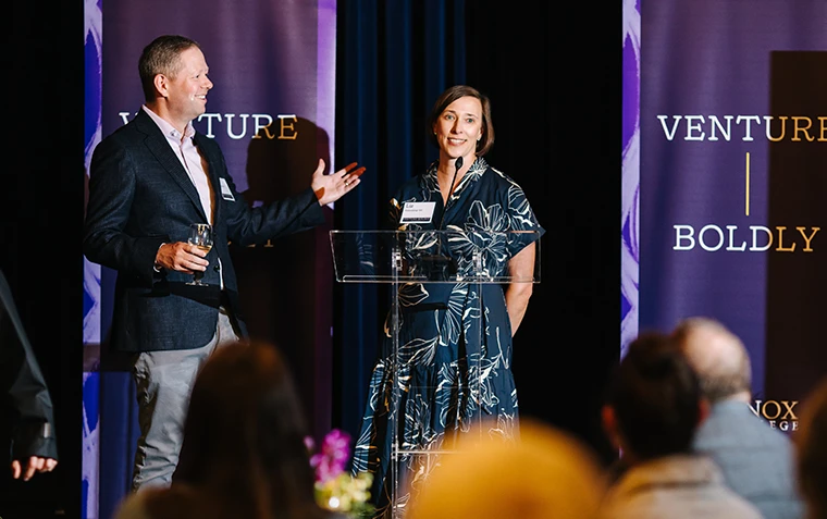 A man and woman speak on stage, flanked by purple Venture Boldly banners