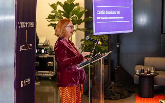 A woman stands at a podium; a Venture Boldly banner is behind her.