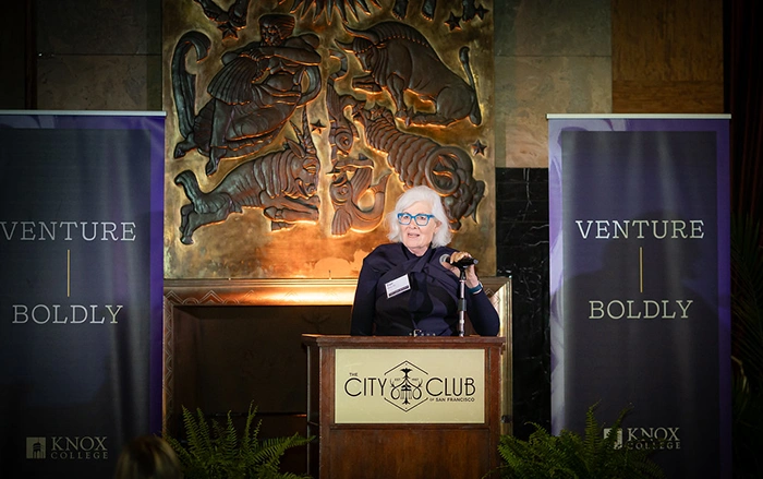 A woman holds a microphone at a podium, standing between Venture Boldly banners.